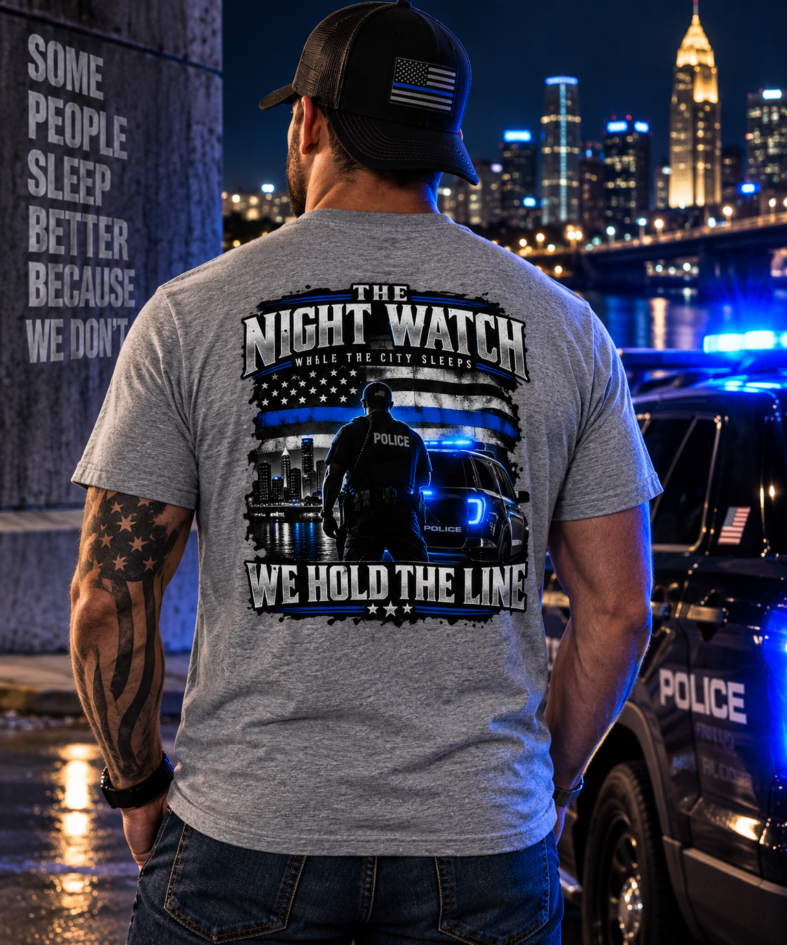 A man viewed from behind wearing a heather gray t-shirt with a large black and blue graphic on the back that reads 'THE NIGHT WATCH WHERE THE CITY SLEEPS WE HOLD THE LINE' with an American flag and police officer design. He is wearing a black baseball cap with an American flag patch and standing next to a police car with blue lights on in front of a city skyline at night.