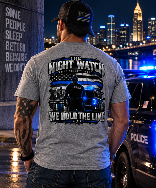 A man viewed from behind wearing a heather gray t-shirt with a large black and blue graphic on the back that reads 'THE NIGHT WATCH WHERE THE CITY SLEEPS WE HOLD THE LINE' with an American flag and police officer design. He is wearing a black baseball cap with an American flag patch and standing next to a police car with blue lights on in front of a city skyline at night.